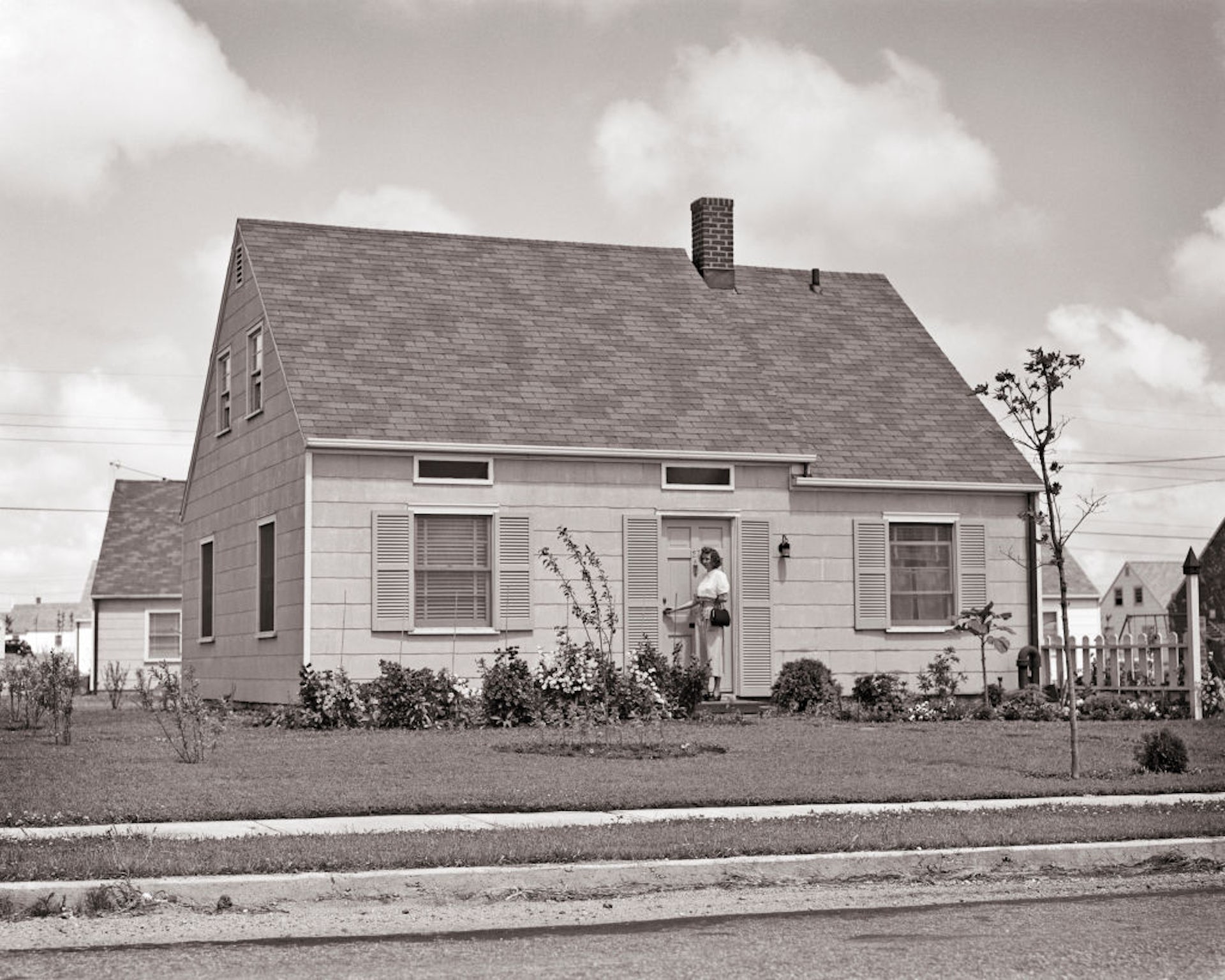 Fotografie alb-negru cu o femeie care sta in afara casei ei suburbane, cu un gazon perfect ingrijit.